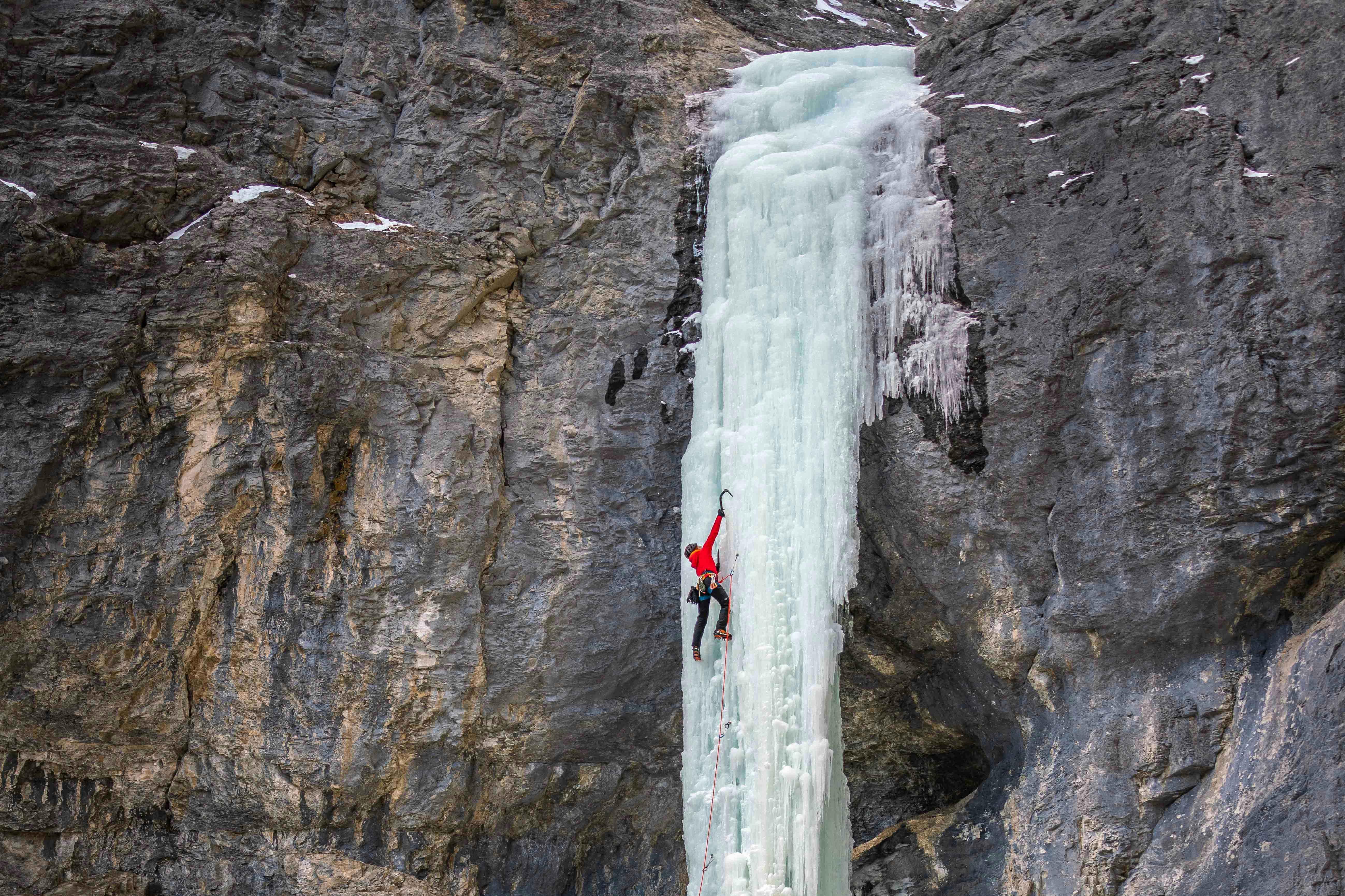 An ice climber reaches up with their axe while climbing a frozen waterfall. The ice is surrounded by large cliffs.