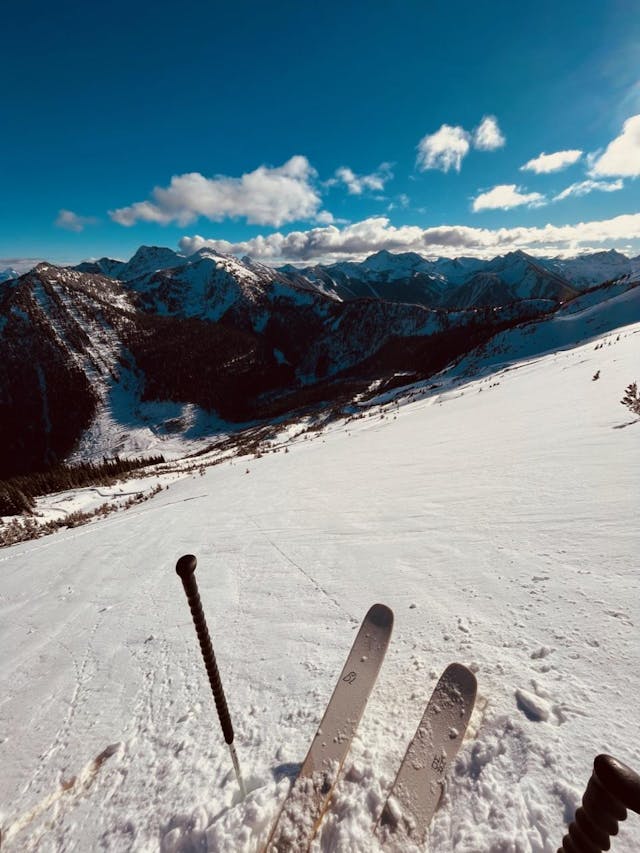A pair of ski tips and a pole are in the front of the photo. Below them, there is a slope with thin and crusty-looking snow. There are some pinwheels and chunks of crust visible below the ski tips.