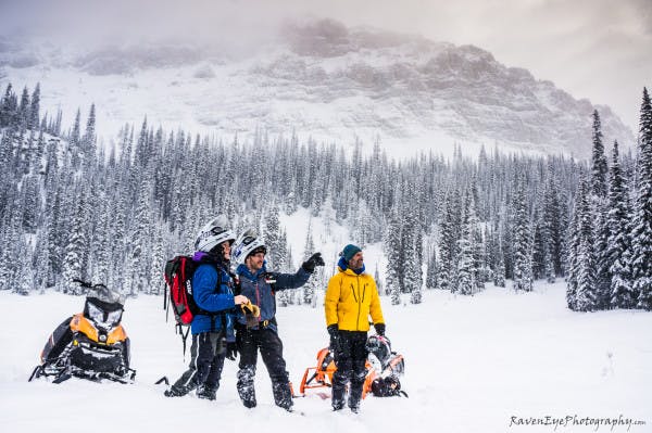 A group of sledders stand in a flat meadow looking at terrain off camera to the right. One is pointing out a feature, while the others look where he is gesturing.