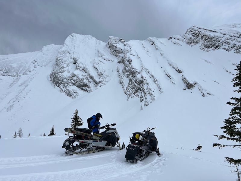 A field team member beside two Ski-Doo snowmobiles in front of an alpine ridge feature