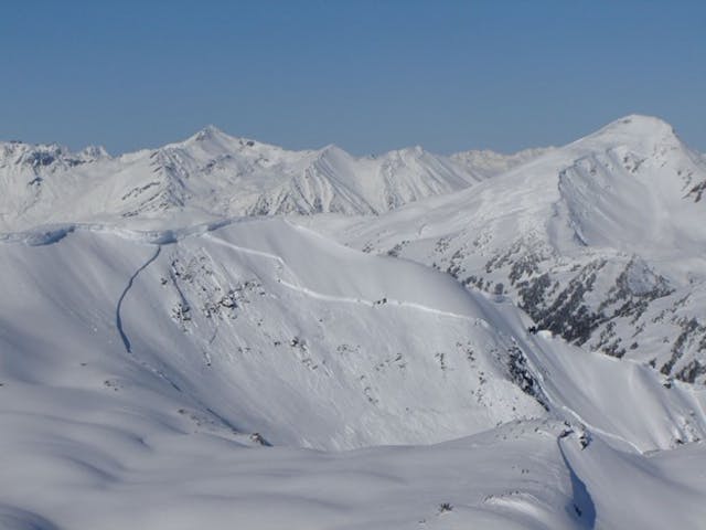 A large avalanche covers much of a slope in bright sunlight. Above the crown, a cornice is still visible. 