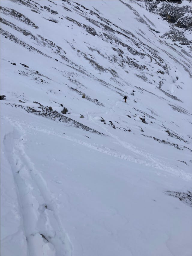 A skier makes their way across a slope with shallow snow coverage. Rocks and scrub are still visible through the snow. 