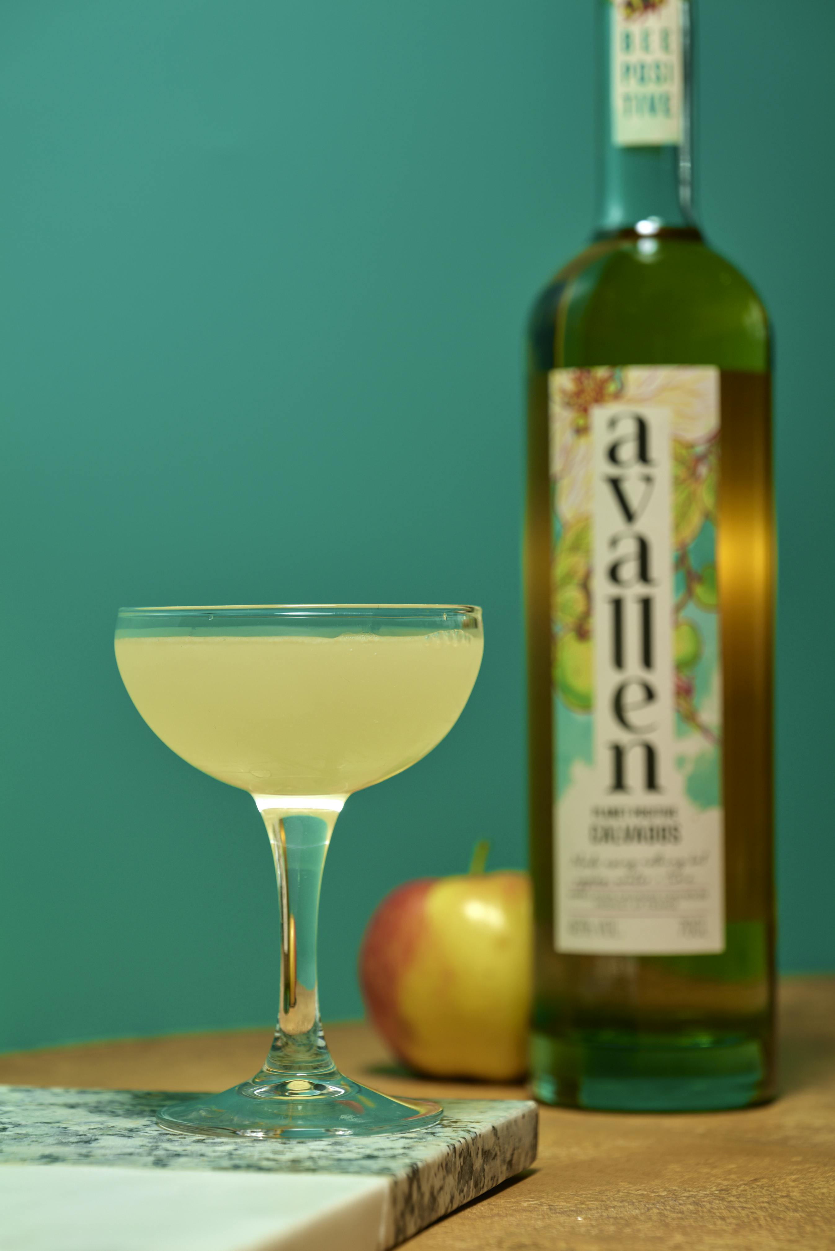 An appletini cocktail in a coupe glass with a bottle of Avallen calvados and an apple
