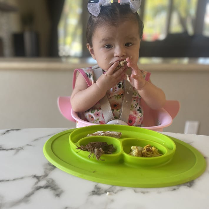 Photograph of baby seated in high chair with ezpz suction Mini Mat of food in front and she is brining food to her mouth