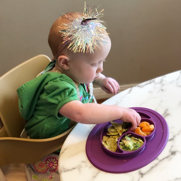 Photograph of baby seated in a high chair reaching into a suction ezpz Mini Mat to pick up food to self-feed