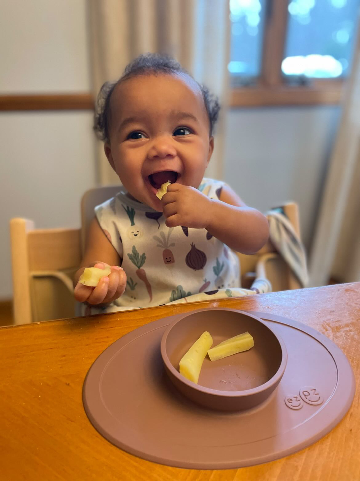 Photograph of baby seated iun high chair with strips of food in an ezpz suction Tiny Bowl smiling and bringing food to her mouth