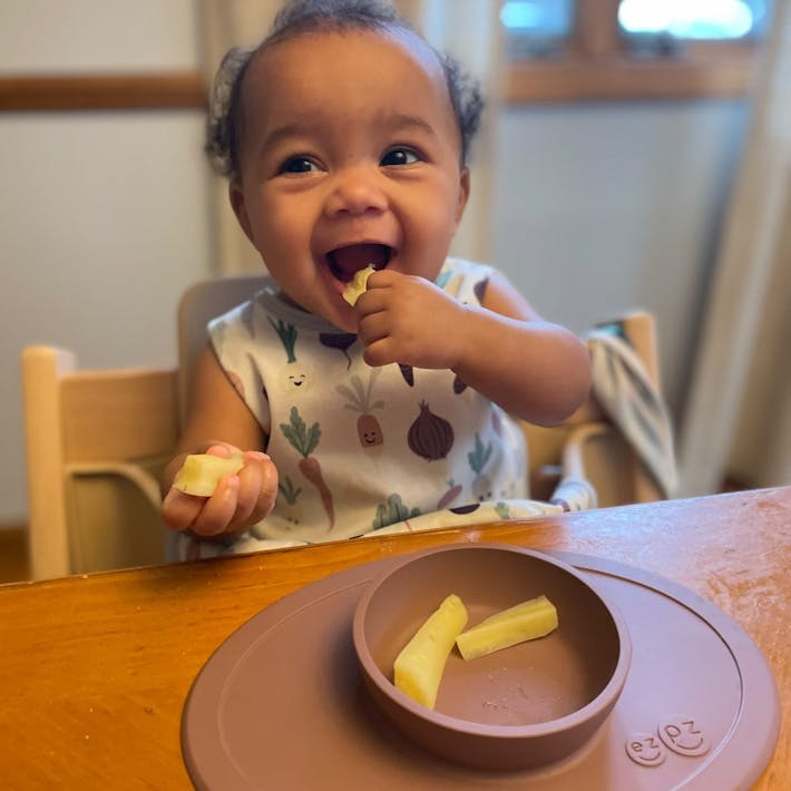 Photograph of baby seated iun high chair with strips of food in an ezpz suction Tiny Bowl smiling and bringing food to her mouth