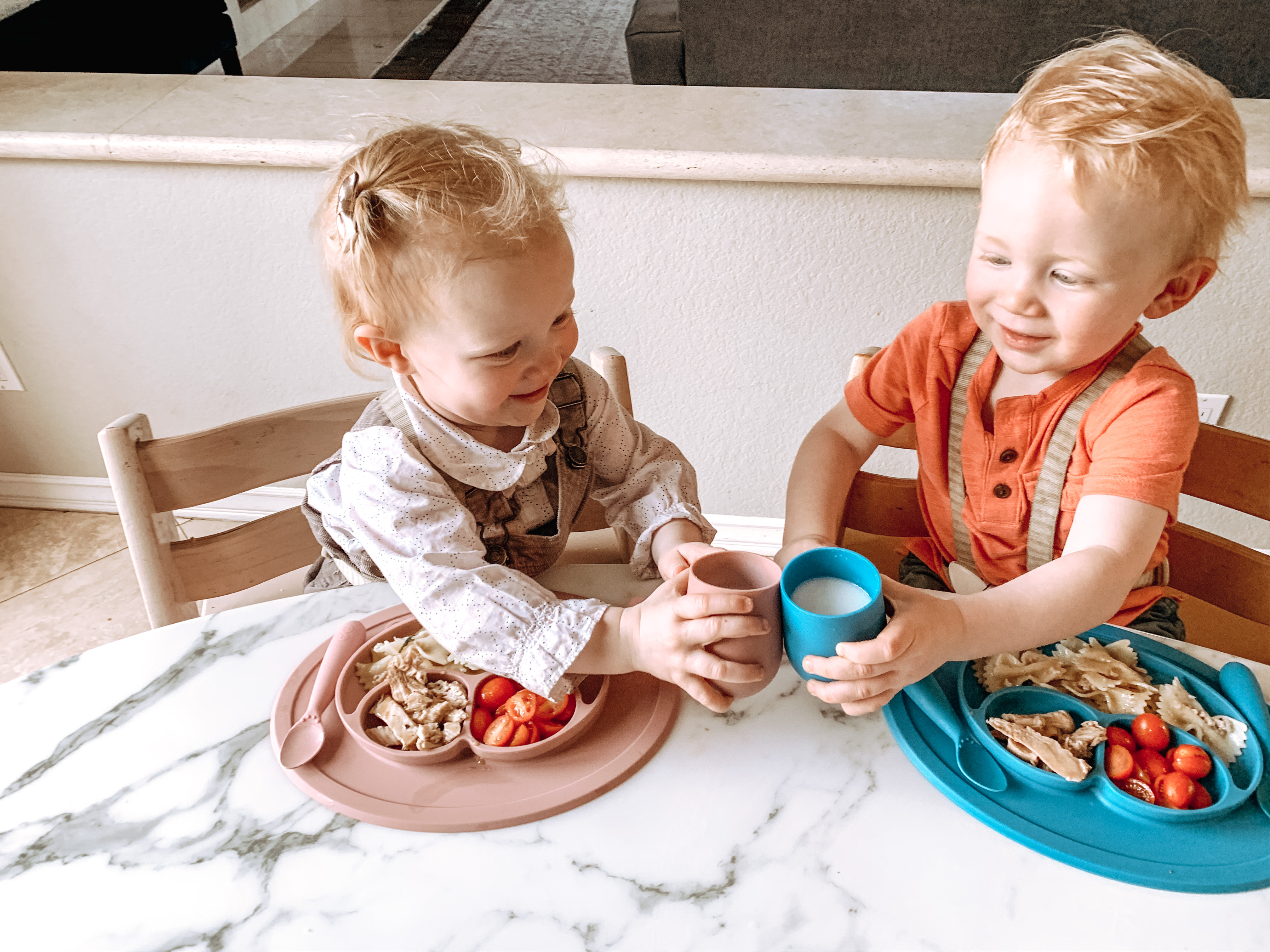 Two toddlers seated in high chairs doing "cheers" with ezpz Mini Cups with ezpz Mini Mats of food in the foreground