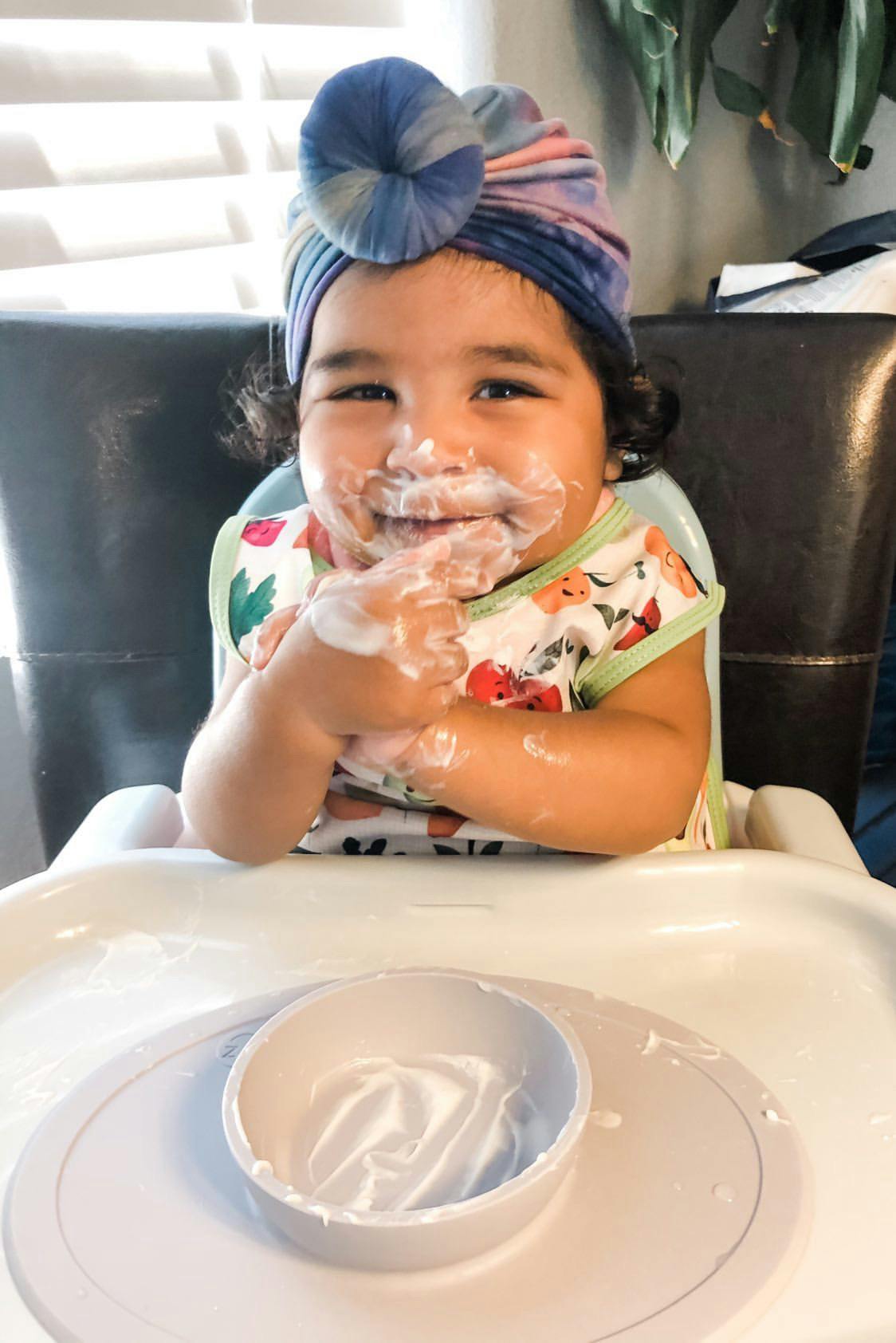Baby seated in high chair with an empty bowl of yogurt in the foreground, messy yogurt smile face
