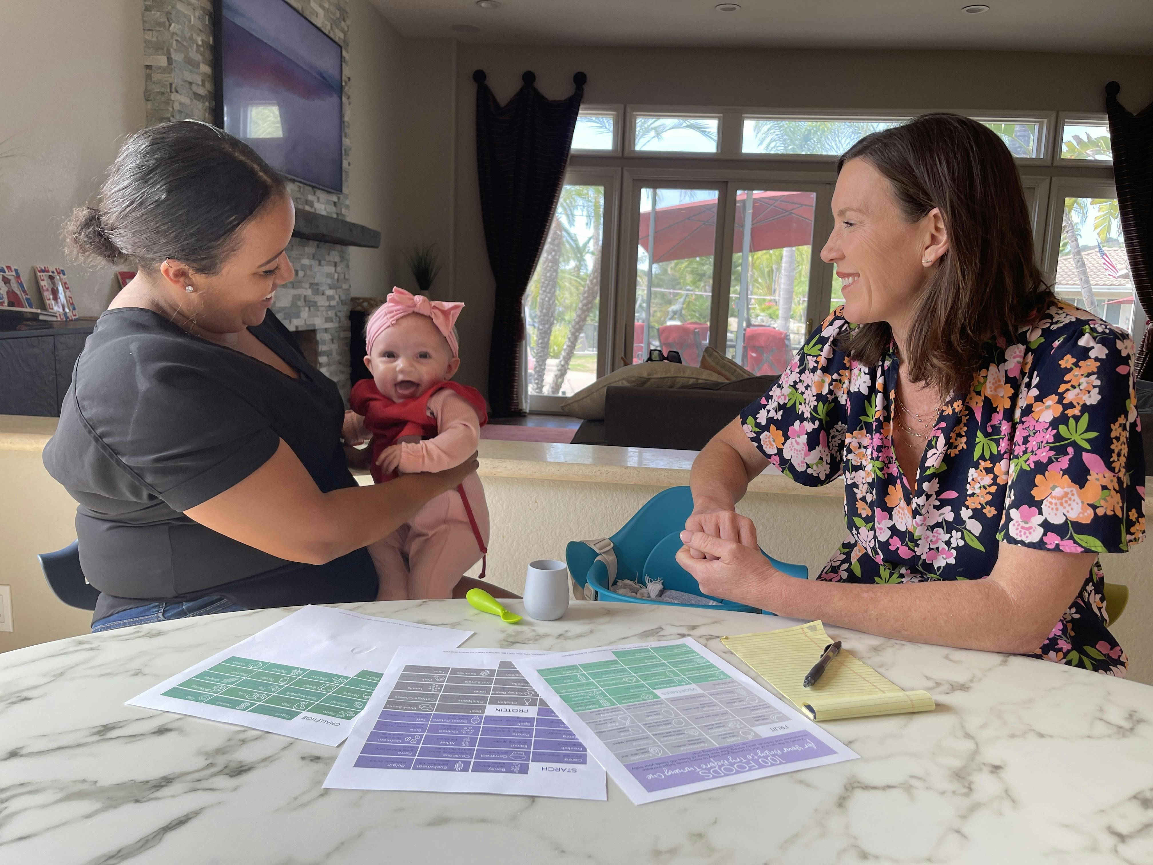 Photograph of mom holding a baby seated at table sitting next to dietitian Katie Ferraro with the 100 FIRST FOODS list spread out on the table in the foreground