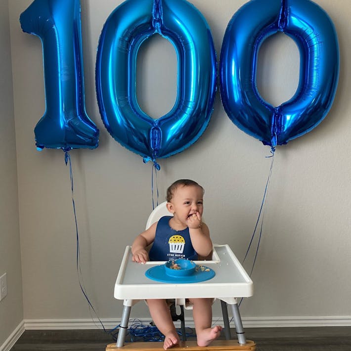 Baby seated in high hair with blue 1-0-0 balloons behind him indicating his 100th First Foods celebration