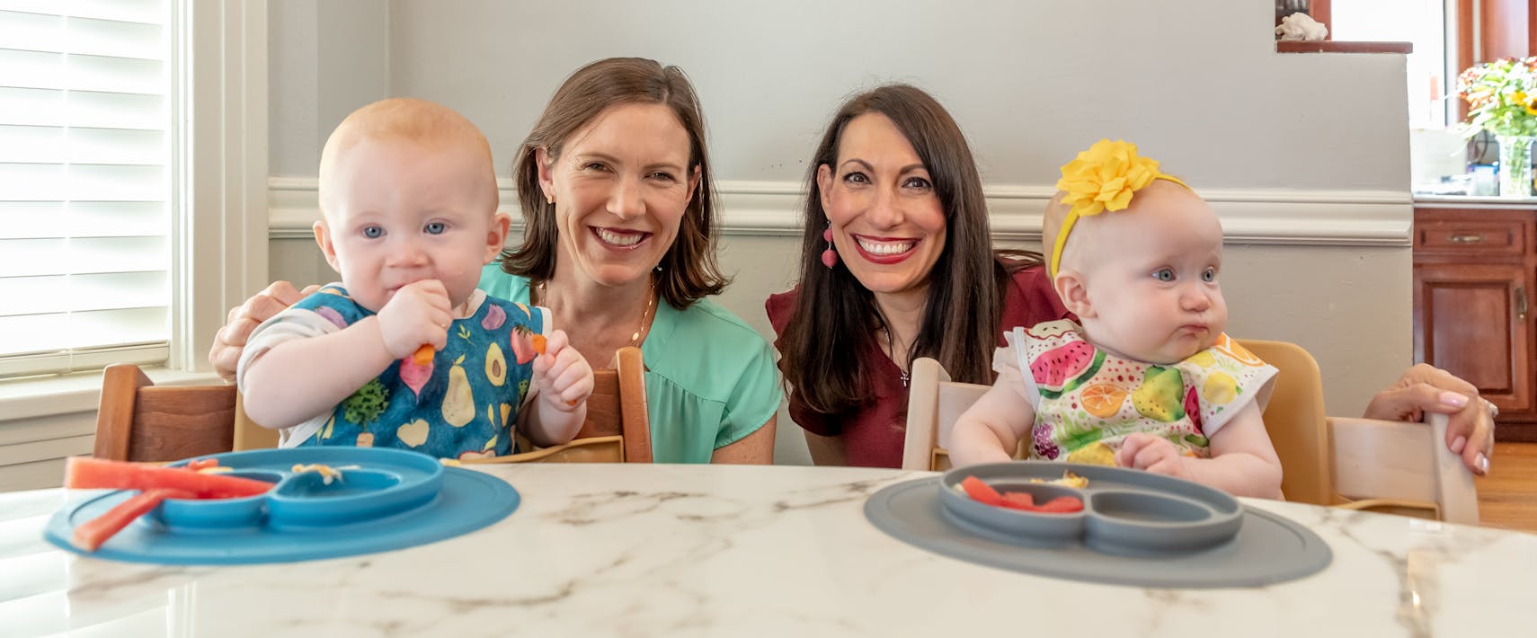 Feeding experts Katie Ferraro and Dawn Winkelmann crouched down together between two babies seated in high chairs who are eating food out of ezpz suction Mini Mats.