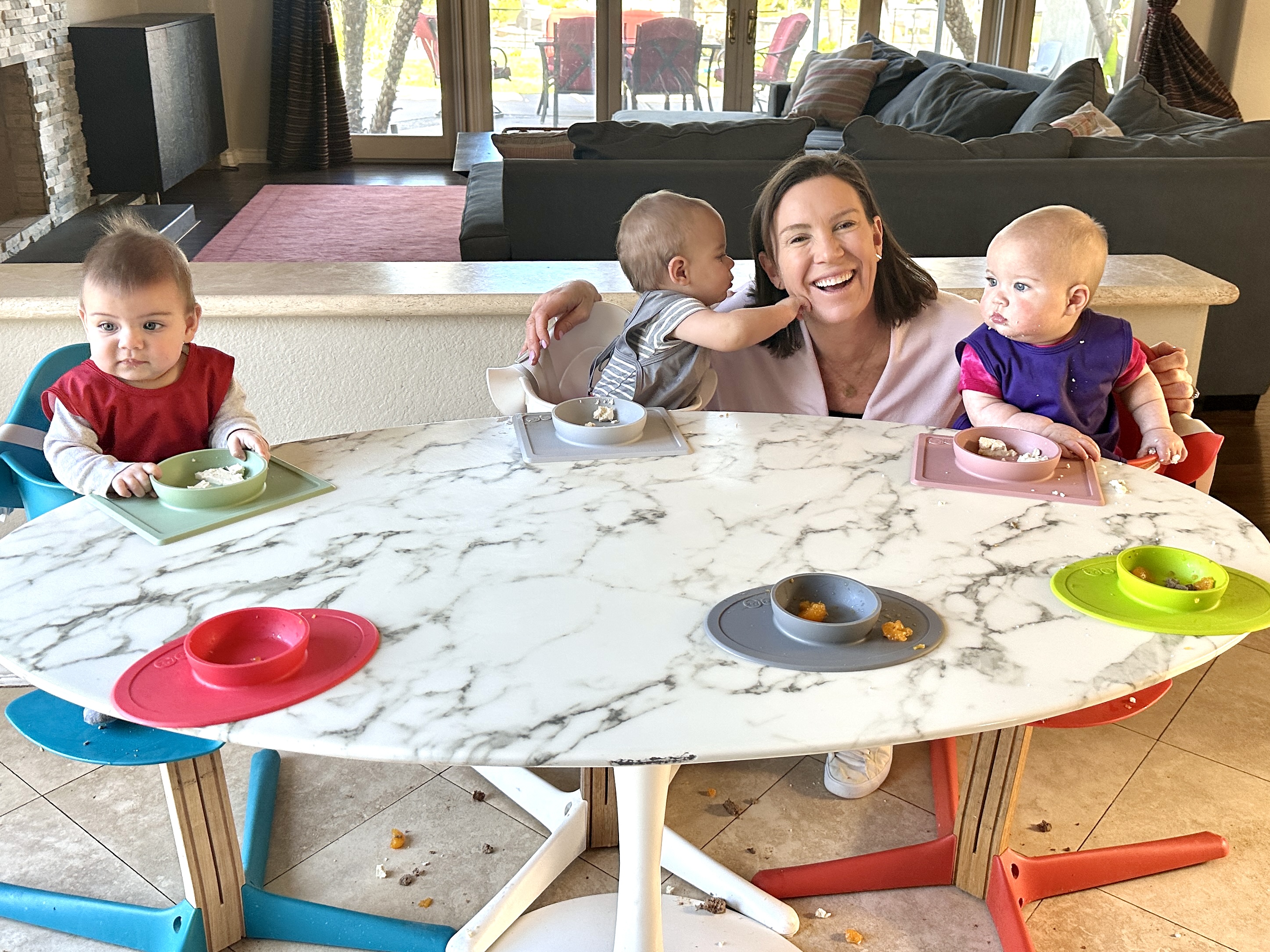 Photograph of Katie Ferraro crouched between two babies who are in high chairs and one is pushing her face while the other is touching an ezpz suction Mini Bowl with food in it.