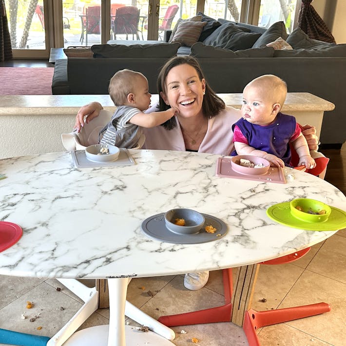 Photograph of Katie Ferraro crouched between two babies who are in high chairs and one is pushing her face while the other is touching an ezpz suction Mini Bowl with food in it.