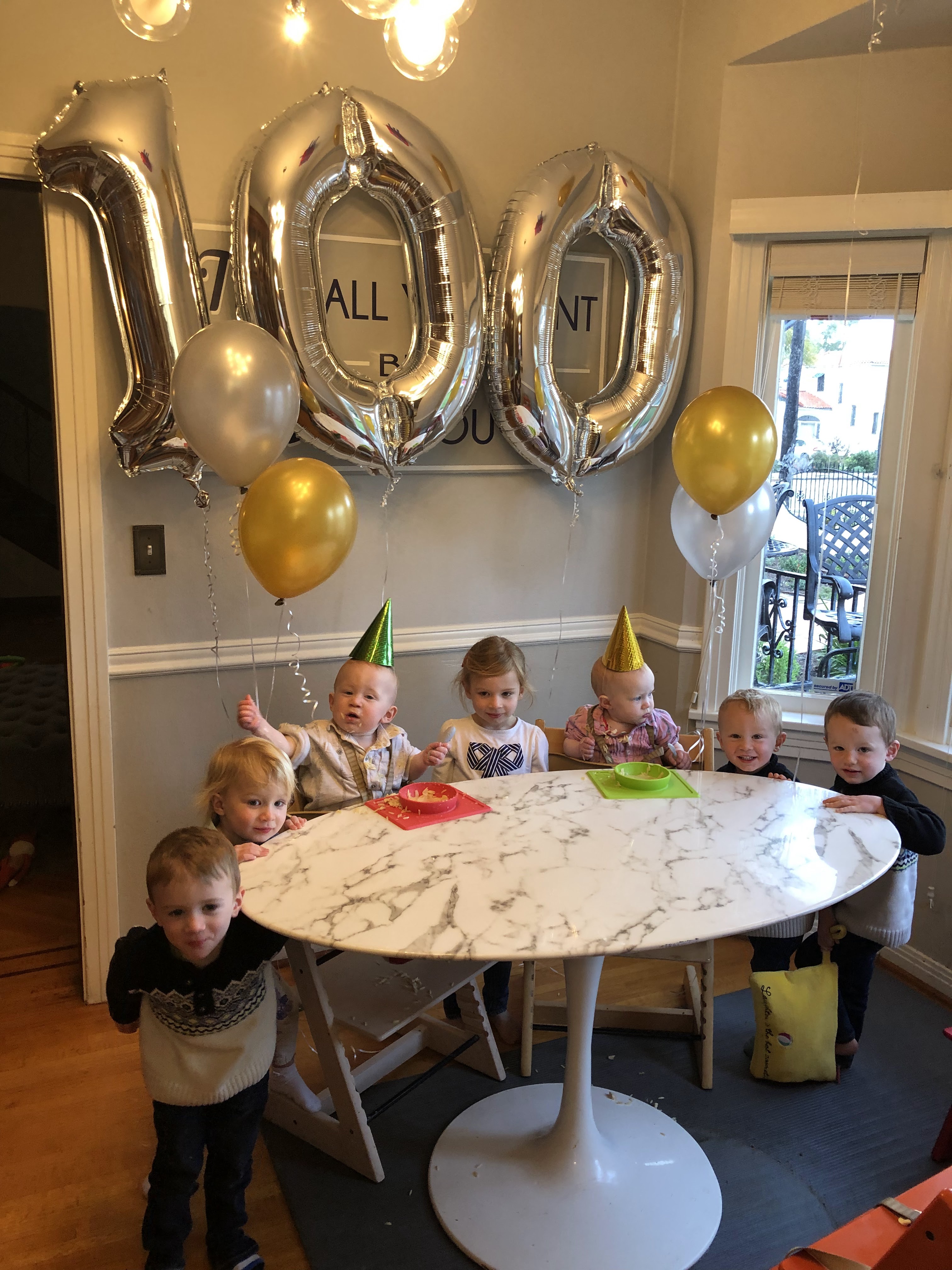 Baby twins sitting in high chair with party hats on and balloons in background that say 100. Celebrating the babies' 100th First Food along with 5 other standing siblings at the same table.