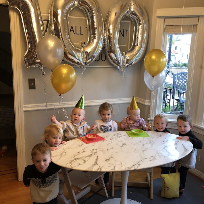 Baby twins sitting in high chair with party hats on and balloons in background that say 100. Celebrating the babies' 100th First Food along with 5 other standing siblings at the same table.