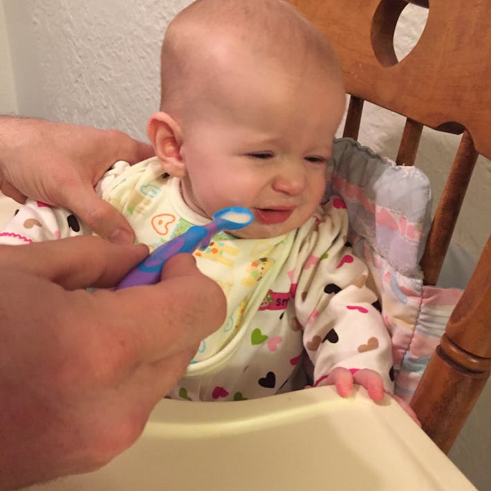 Baby in high chair turning her face away from a spoon of food being presented to her mouth by an adult hand