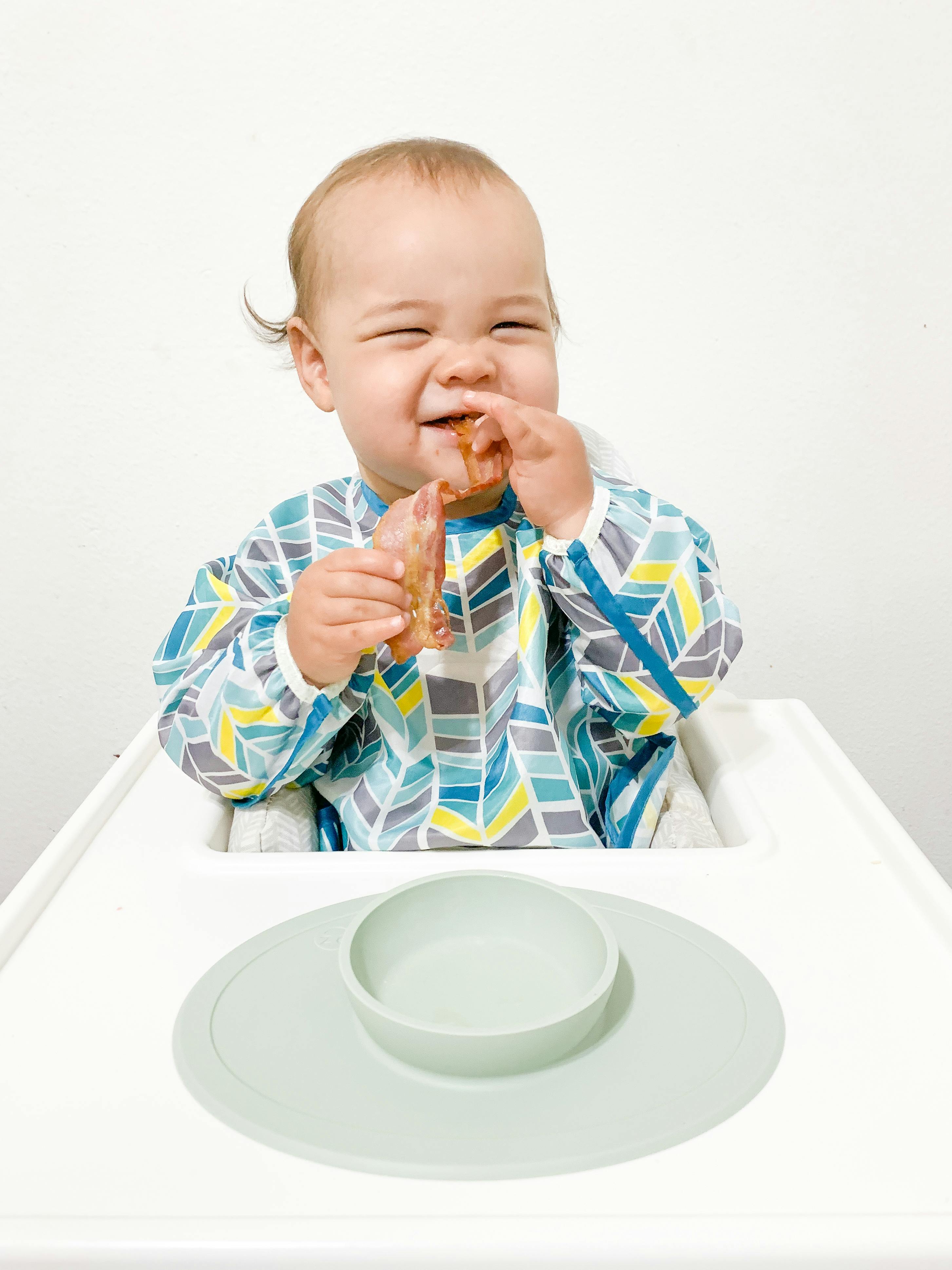 Photograph of a baby seated in a high chair bringing a strip of bacon from an ezpz Tiny Bowl to his mouth using both hands and smiling.