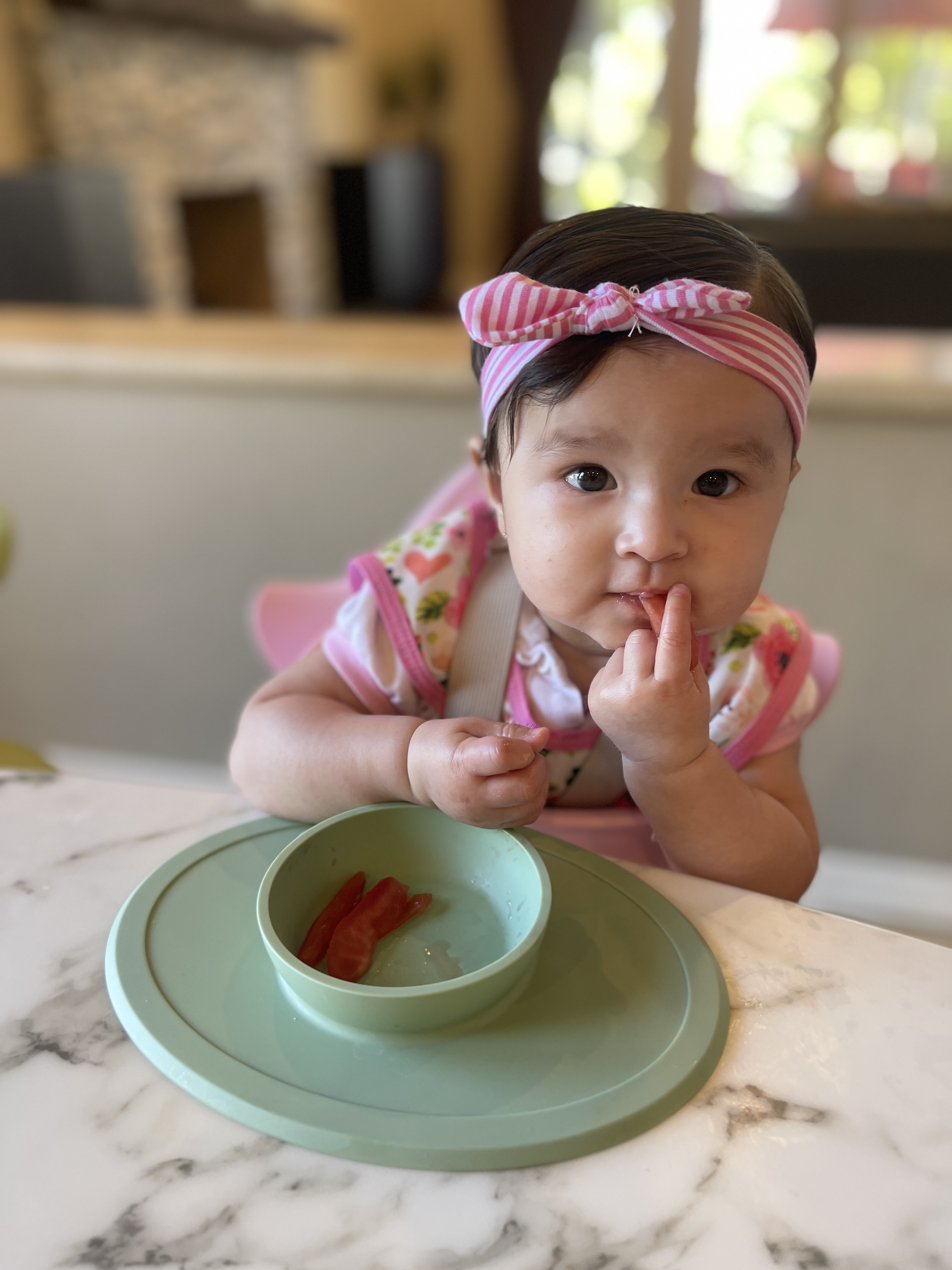 Photograph of baby seated in a high chair leaning into an ezpz suction Tiny Bowl with plum strips in the bowl and bringing some food to her mouth