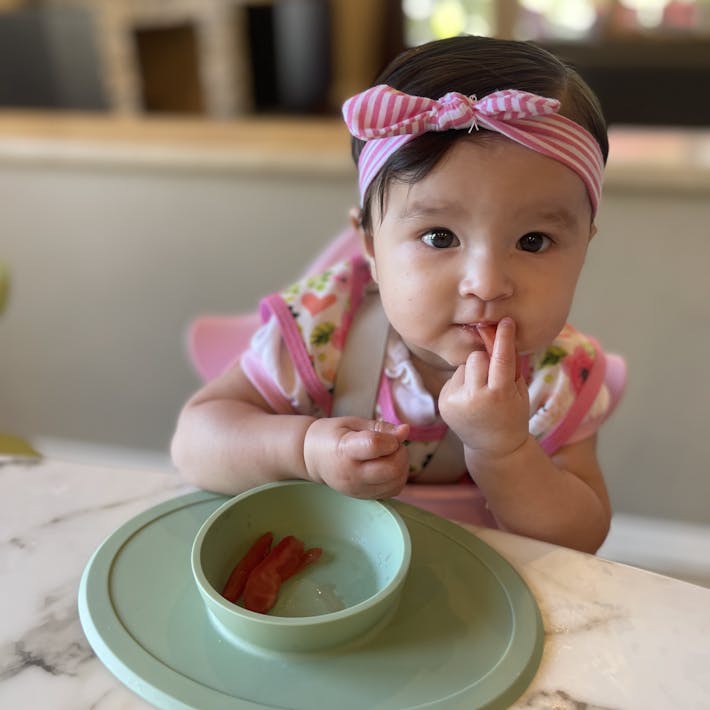 Photograph of baby seated in a high chair leaning into an ezpz suction Tiny Bowl with plum strips in the bowl and bringing some food to her mouth