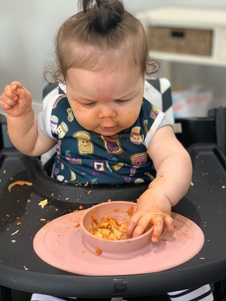 Photograph of baby seated in a high chair reaching for food using her pincer grasp to pick up food out of an ezpz Tiny Bowl