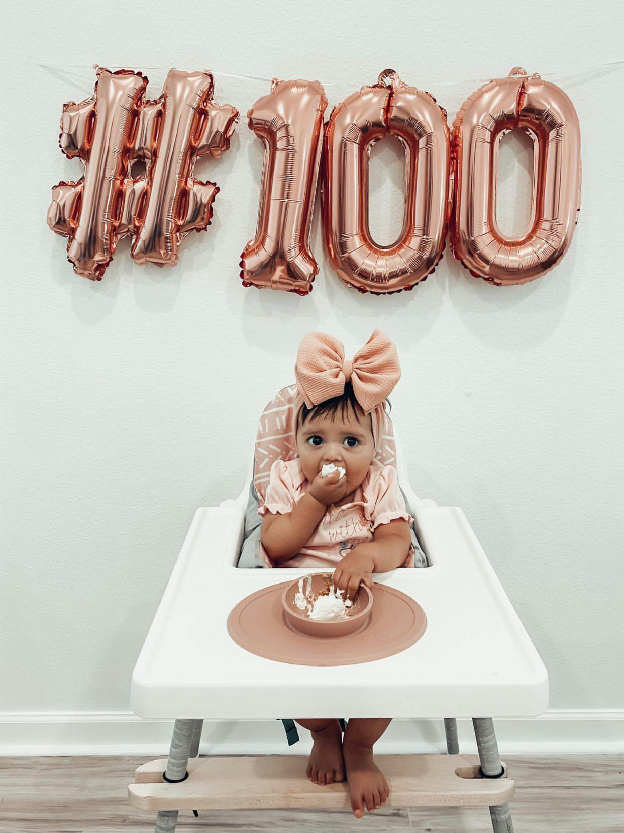 Photograph f a baby seated in Ikea Antelop high chair with 3rd party footrest and eating food out of an ezpz Tiny Bowl. Pictured in the back are balloons forming the # 1 0 0 indicating the baby's 100th first food.