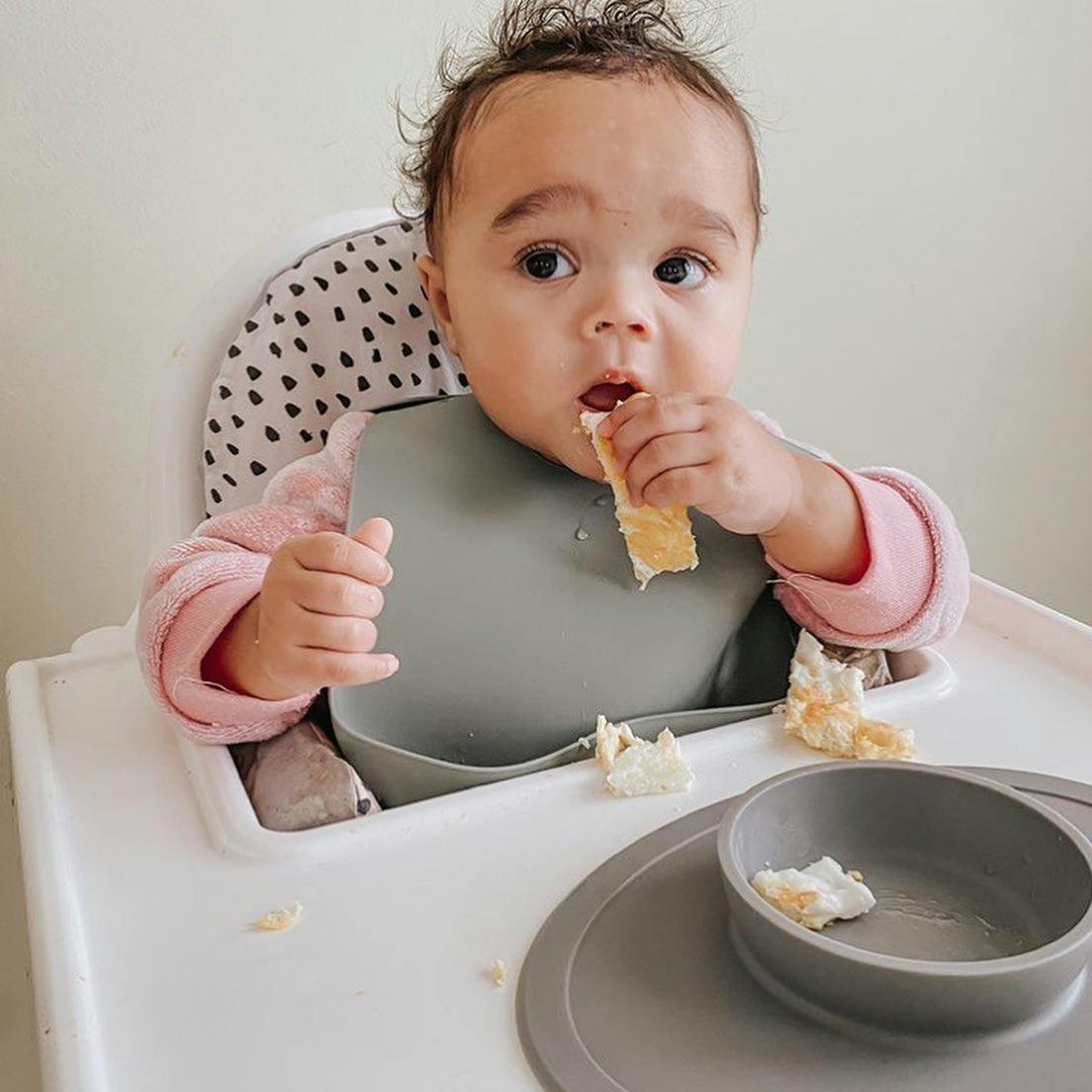 Photograph of a baby in a high chair bringing a trip of fried egg from an ezpz Tiny Bowl to her mouth with her left hand.