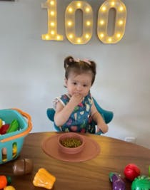 Baby Alma seated in high chair with suction bowl of food in front of her, bringing food to mouth and positioned in front of balloons spelling out 1-0-0