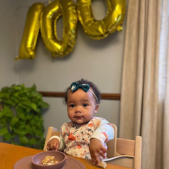 Photograph of baby in her high chair with food in an ezpz Tiny Bowl featured in foreground and balloons in background spelling out 1-0-0 to celebrate the baby's 100th first food before turning one.