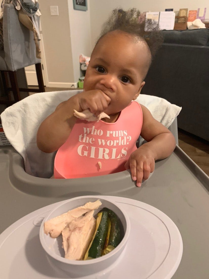 Photograph of baby seated in high chair feeding herself chicken with right hand; pictured in foreground is an ezpz Tiny Bowl with chicken and asparagus.