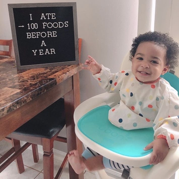 Photograph of baby seated in high chair pointing ot a letter board that says I ATE 100 FOODS BEFORE A YEAR