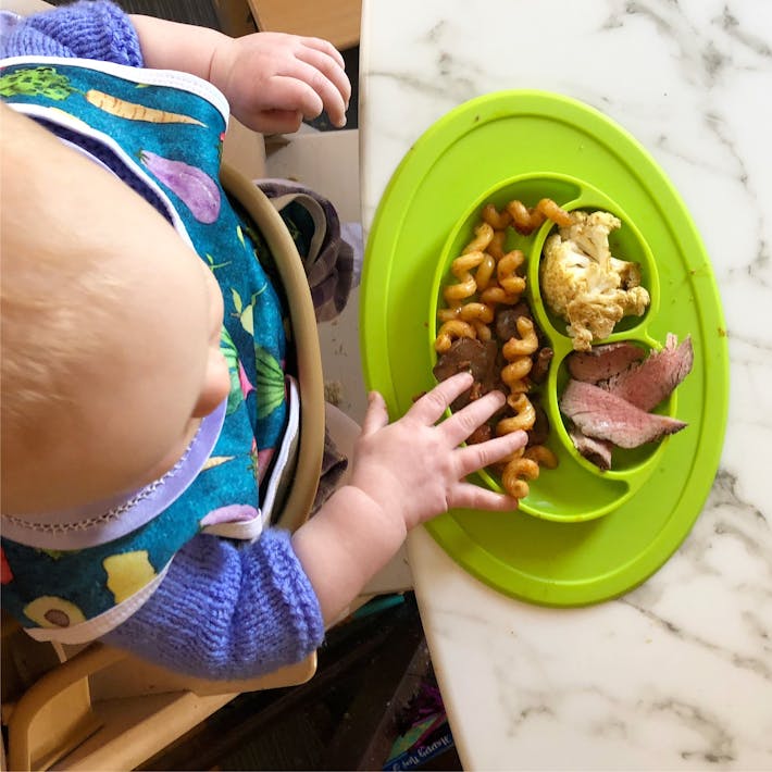 Overhead photograph looking down on baby seated in a high chair with her hand reaching into the ezpz Mini Mat which is filled with food
