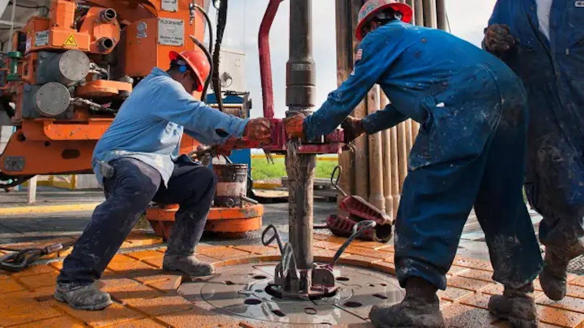 Two workers wearing hard hats handle heavy equipment in an oil production facility