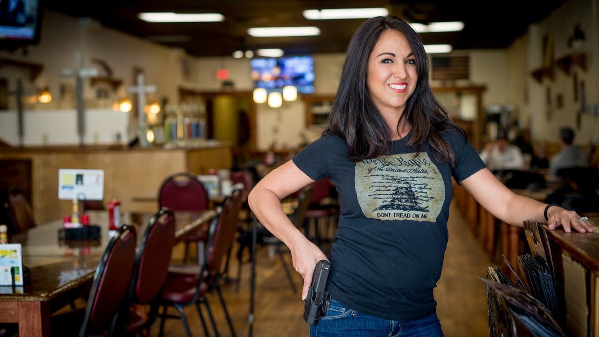 Image of Representative Lauren Boebert inside a tacky, grill-type eatery, grinning, leaning on a host stand with one hand while grabbing a holstered gun with the other. Her trigger finger is extended.
