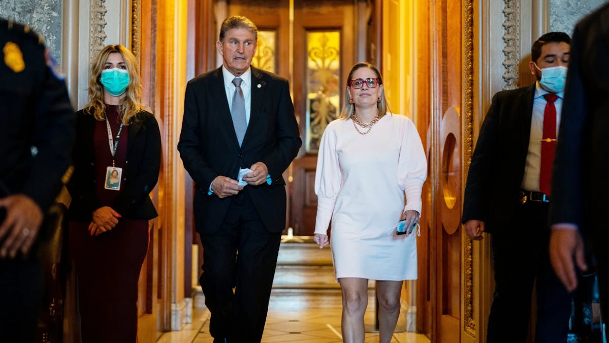 Senators Joe Manchin and Kyrsten Sinema walking together at the US Capitol Building, flanked by aides wearing face masks.