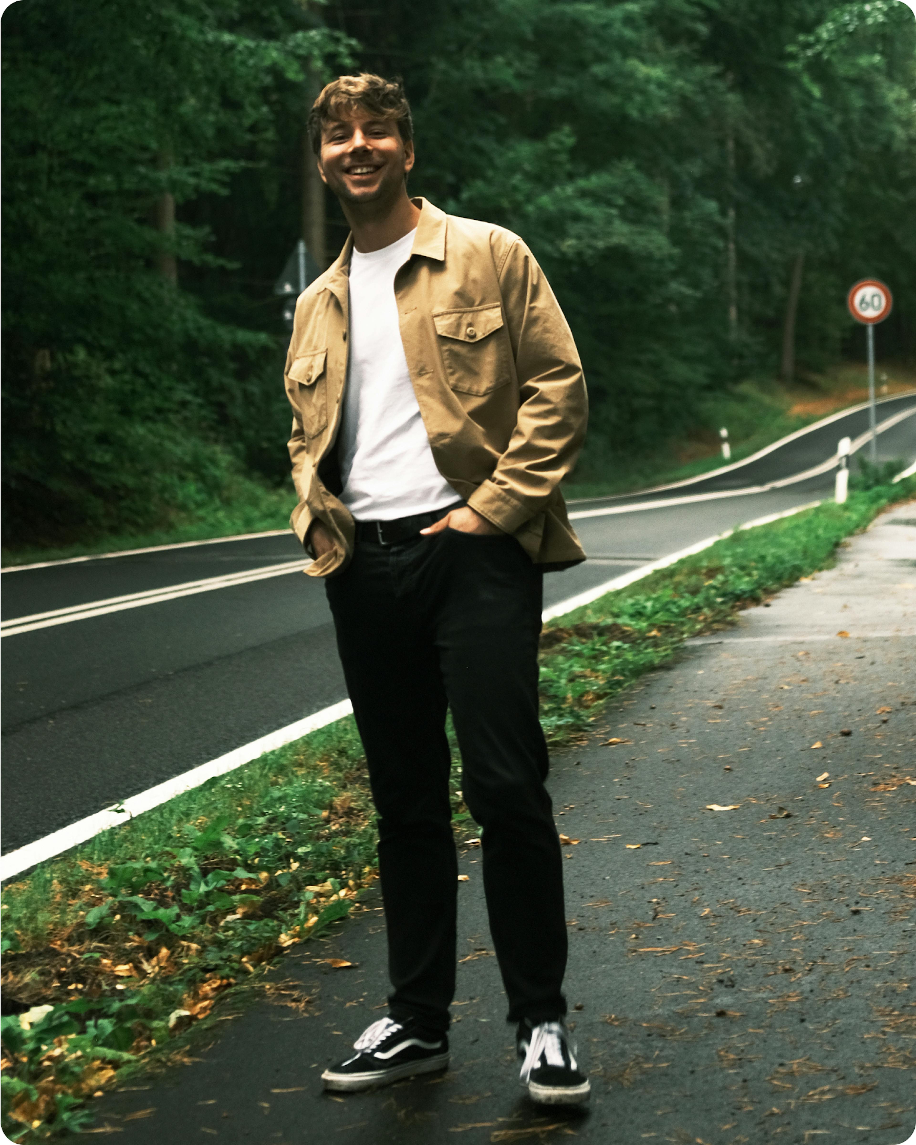 Ein Mann steht am Rand einer Landstraße und lächelt in die Kamera.