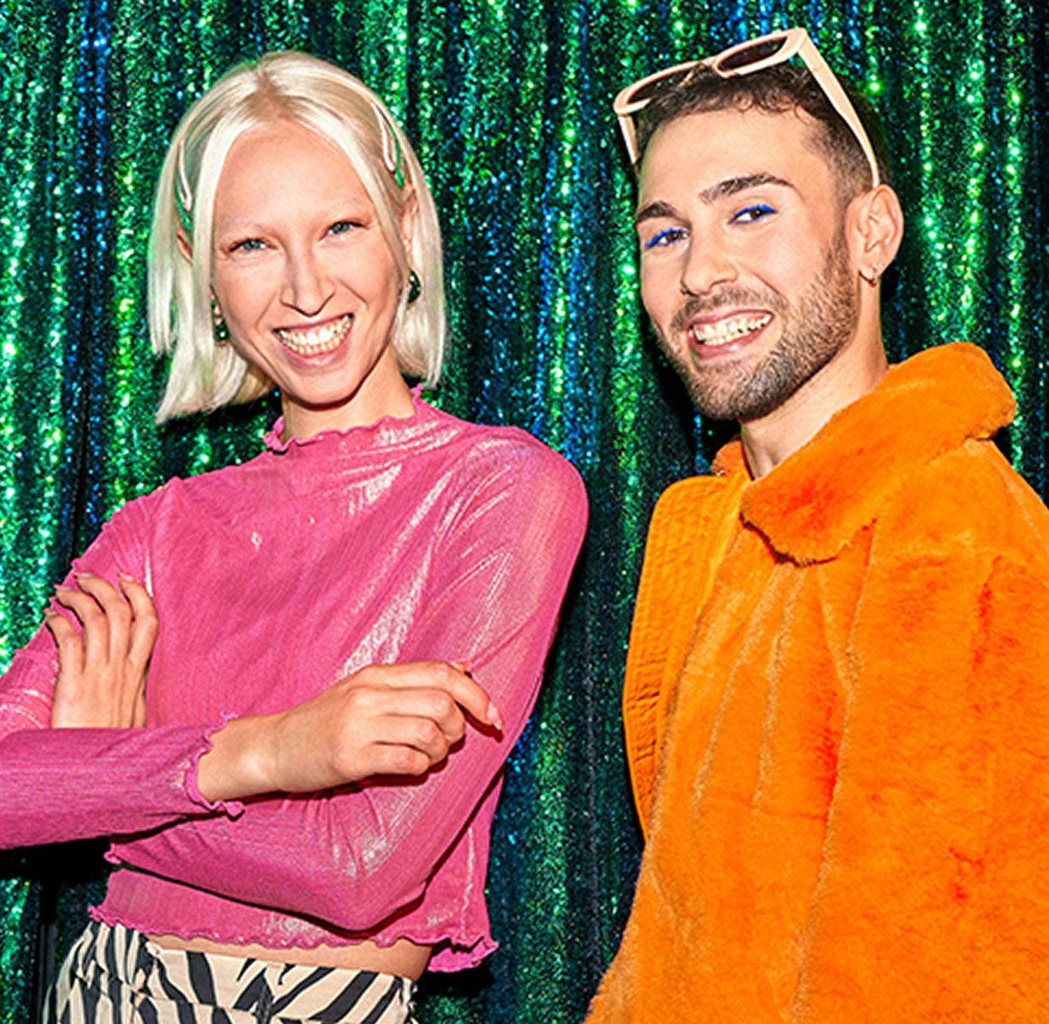 Eine junge Frau und ein junger Mann stehen lachend vor einem grün glitzernden Vorhang.