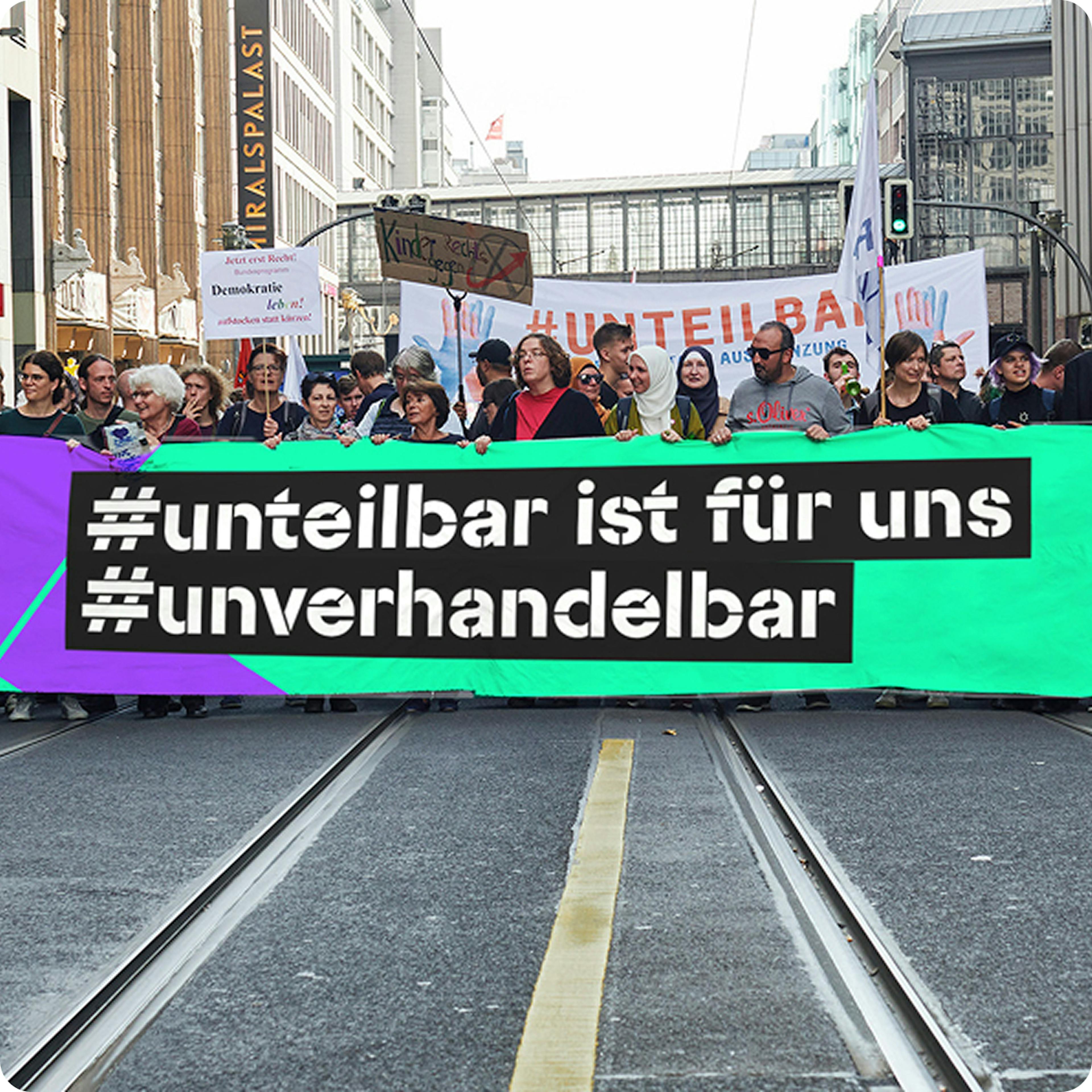 Frontales Bild eines Demonstrationszuges, im Vordergrund großes Banner mit der Aufschrift "#unteilbar ist für uns #unverhandelbar".