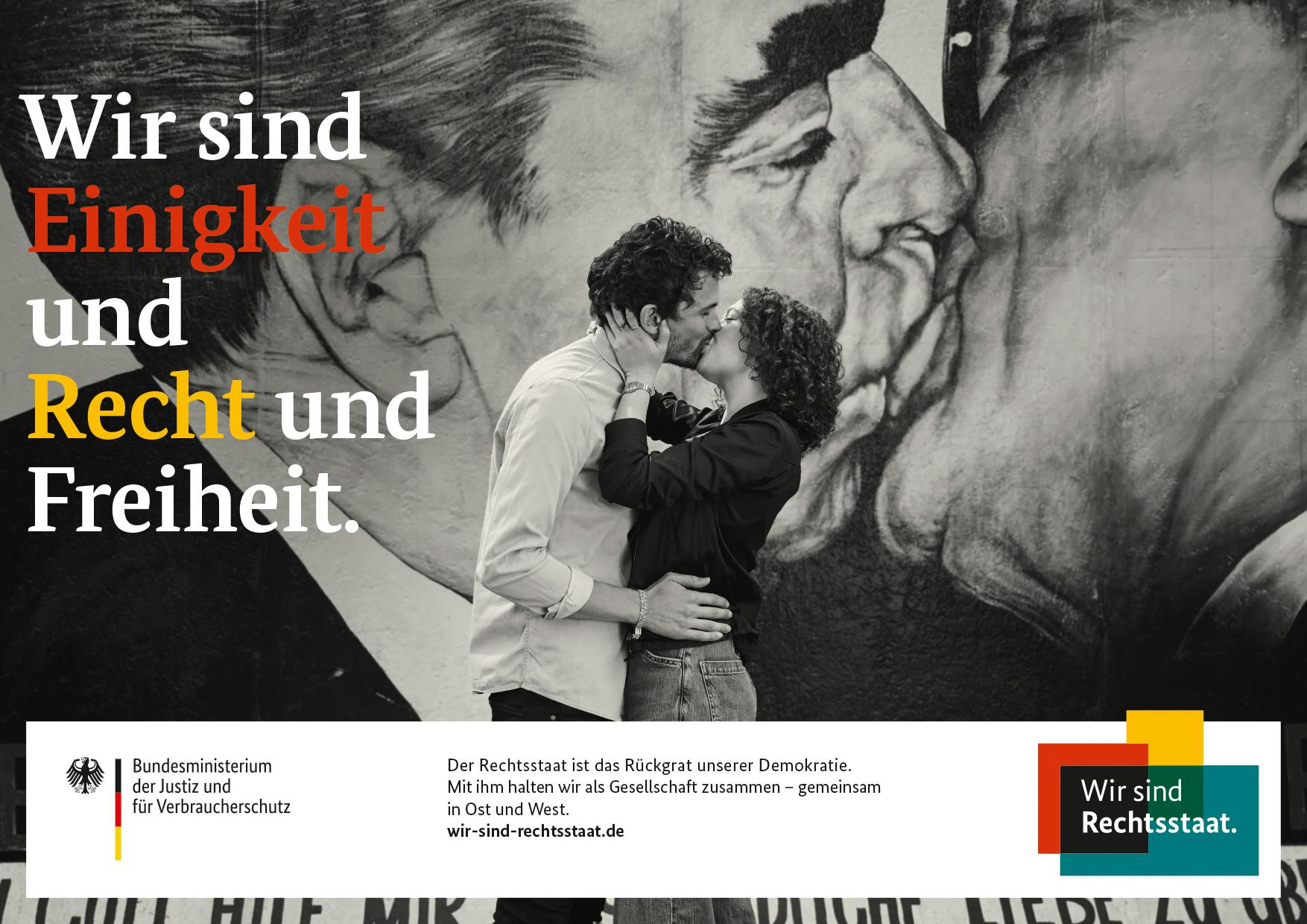 Schwarzweißbild eines sich küssenden Paares vor der Berliner Mauer, daneben Schriftzug "Wir sind Einigkeit und Recht und Freiheit."