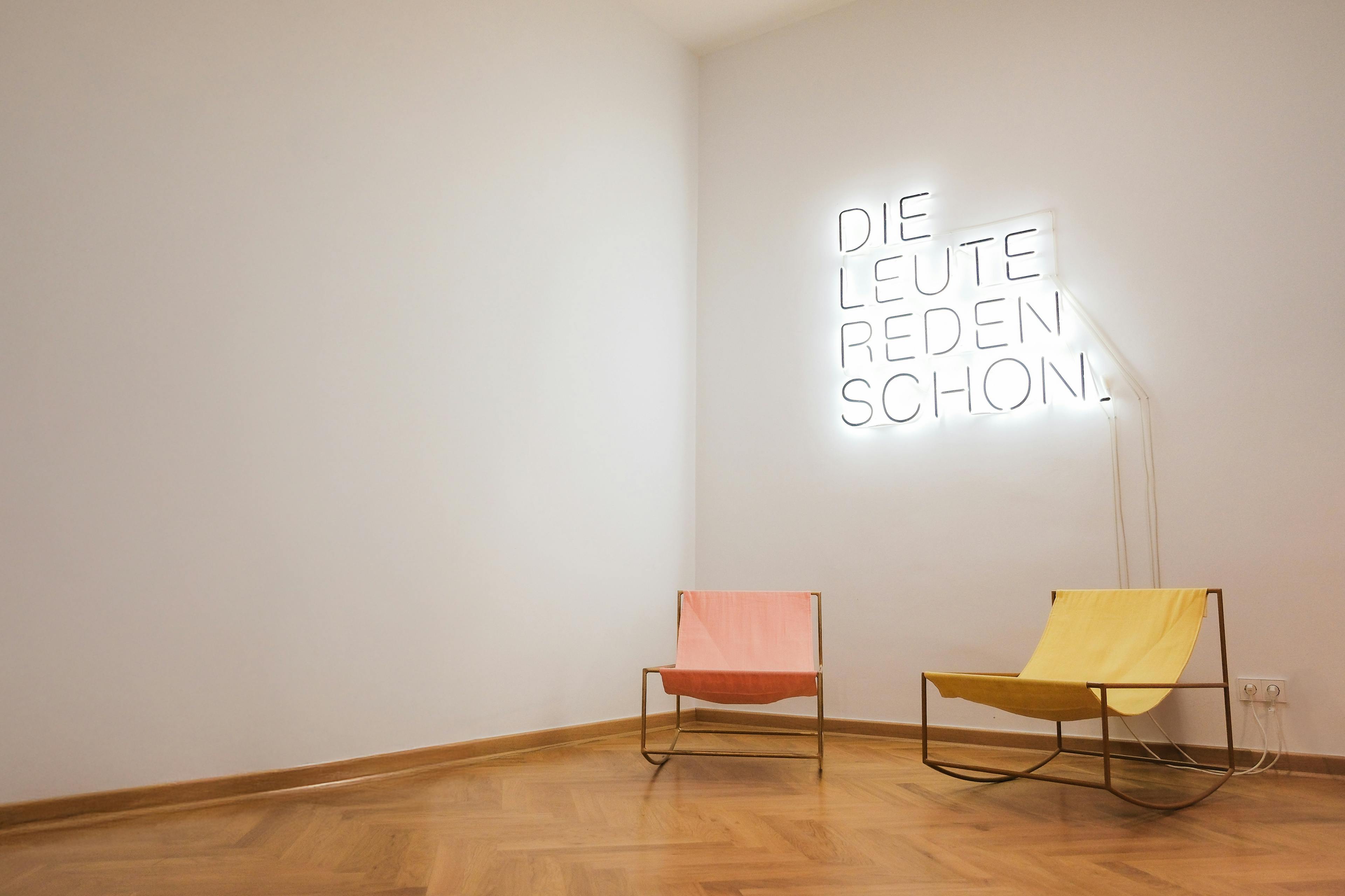 Zwei Liegestühle vor der leuchtenden Neonschrift "Die Leute reden schon".