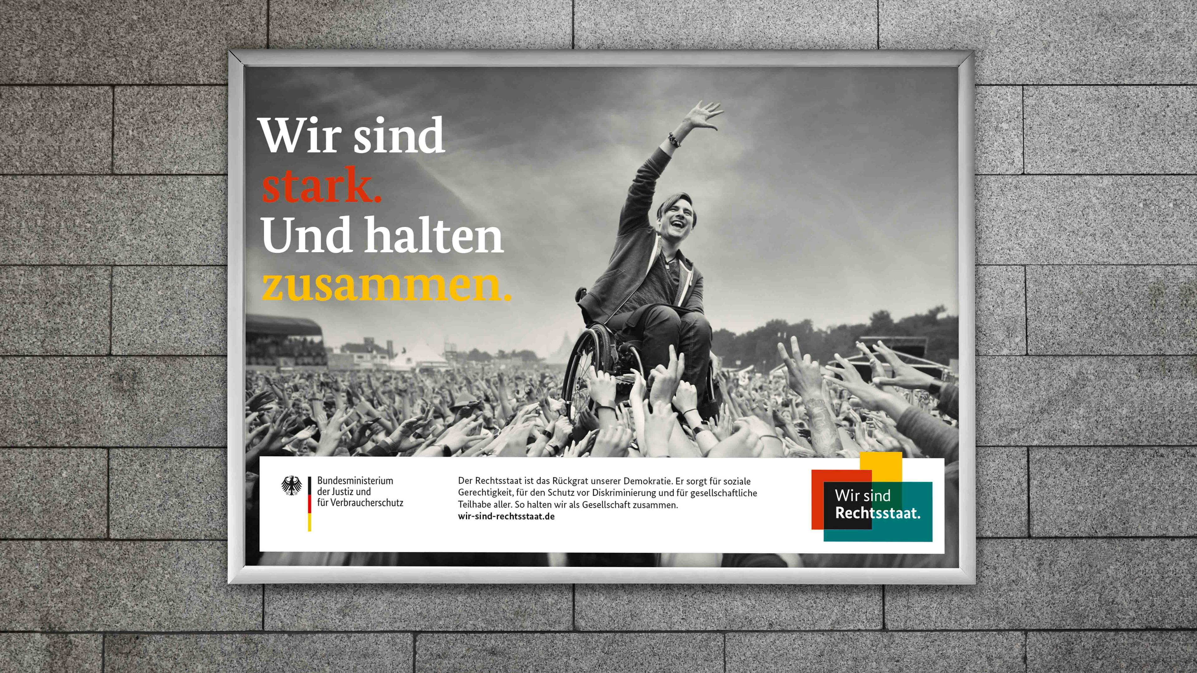 Schwarzweißbild eines Rollstuhlfahrers, der von einer Menge getragen wird. Schriftzug "Wir sind stark. Und halten zusammen."