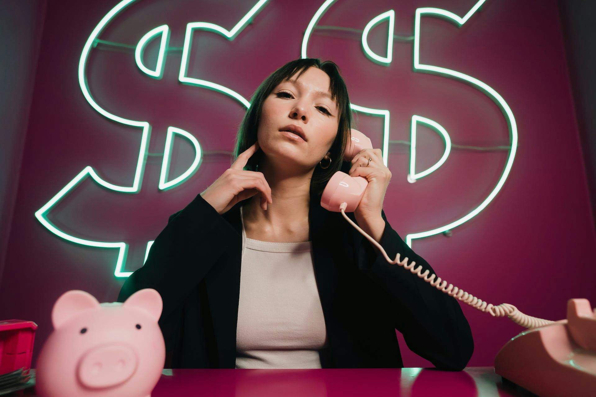 Woman on phone seated in front of neon dollar signs with piggy bank in front of her.