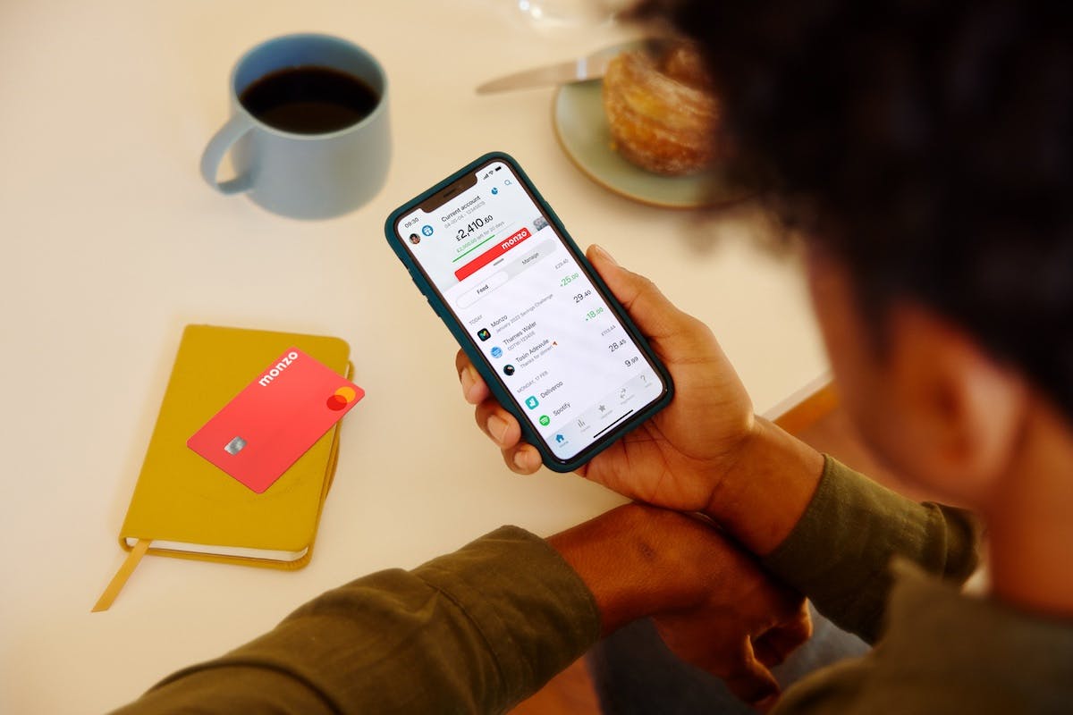 A person holding a mobile phone that shows the Monzo account app. On the table in front of them is a coffee, a croissant, a notebook and a Monzo card.