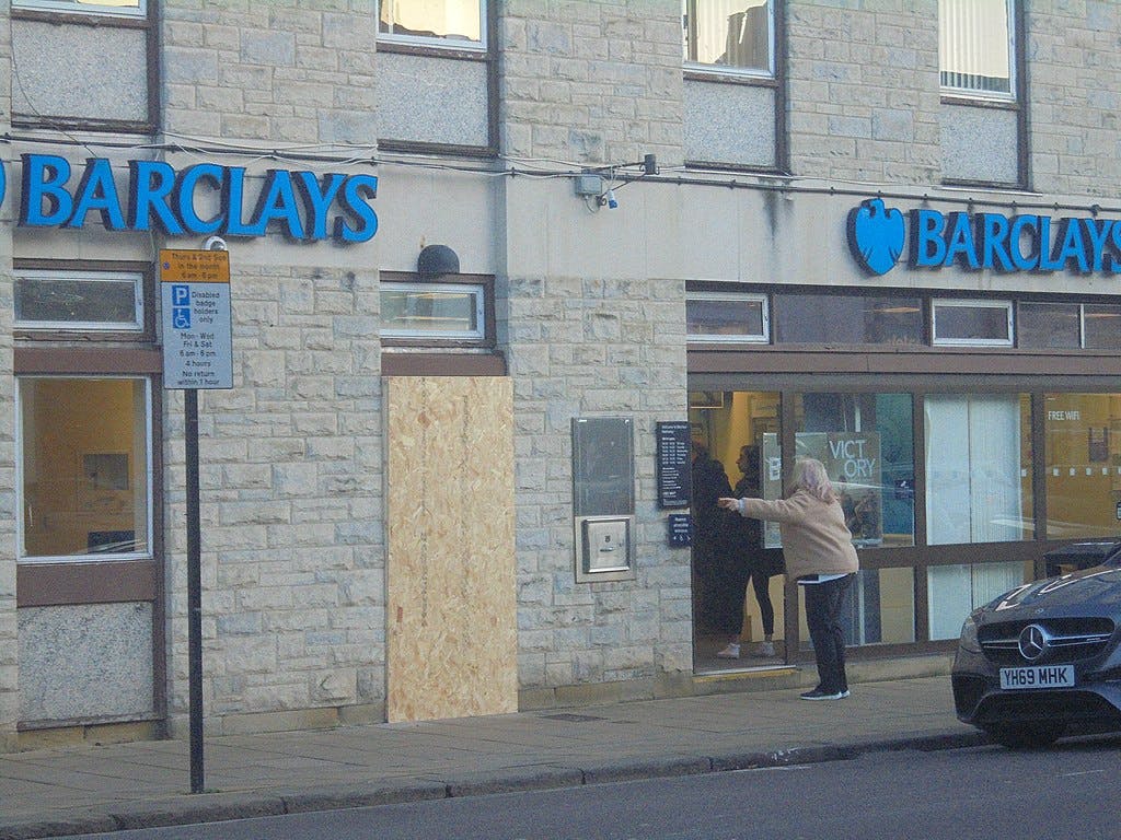 A Barclays bank in Yorkshire that has had its window busted out. An angry woman stands at the door pointing.