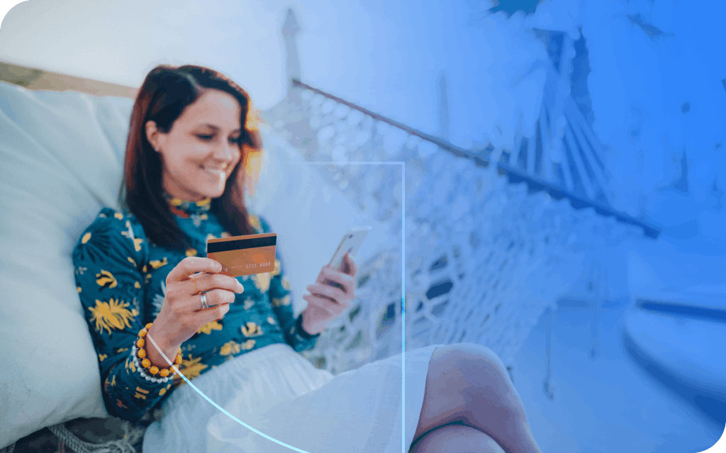 A smiling woman sitting on a hammock holding a credit card in one hand and a smartphone in her other hand