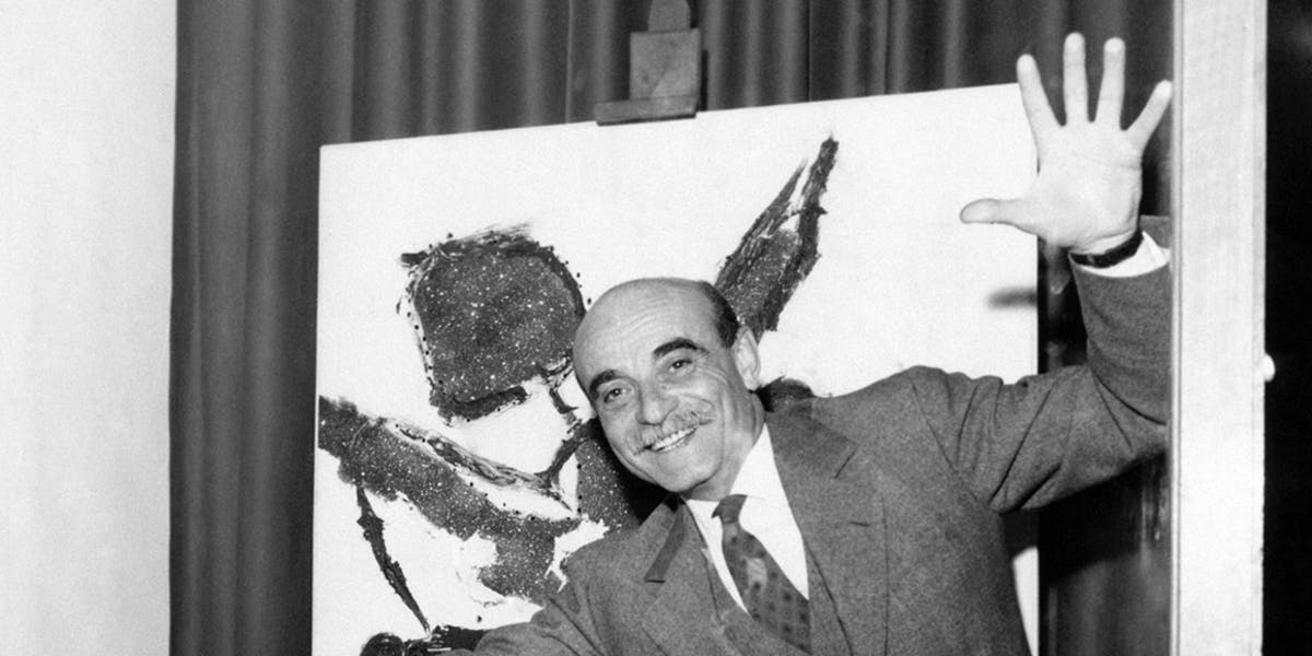 Lucio Fontana jokes in front of a painting in the Galleria del Naviglio. Milan, 1950s. Photo by Mondadori Portfolio via Getty Images