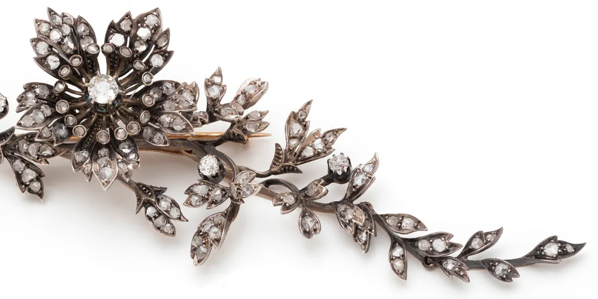 Broche en or 750 et argent 800 millièmes, composée de 2 éléments articulés à décor feuillagé, le plus important centré d'une fleur d'églantine montée en trembleuse, terminée par un ruban noué. Travail français vers 1880, dans un style naturaliste empreint de romantisme. Bijou transformable. Elle est conservée dans son écrin d'origine à la forme en cuir de la maison "MELLERIO dits MELLER", intérieur velours et soie, monogrammé A.L.C. Estimation : 1800 - 2200 euros. (détail)