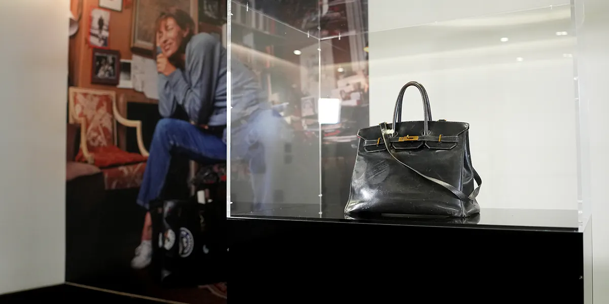 PARIGI, FRANCIA – 3 LUGLIO: La borsa Birkin originale in pelle nera di Jane Birkin è esposta in una teca di vetro trasparente da Sotheby's, con un grande ritratto fotografico dell'attrice nella sua casa sullo sfondo. La borsa consumata e personalizzata è il pezzo forte dell'asta “The Original Birkin” che si terrà il 10 luglio. Fotografata da Sotheby's il 3 luglio 2025 a Parigi, Francia. (Foto di Edward Berthelot/Getty Images) (dettaglio)