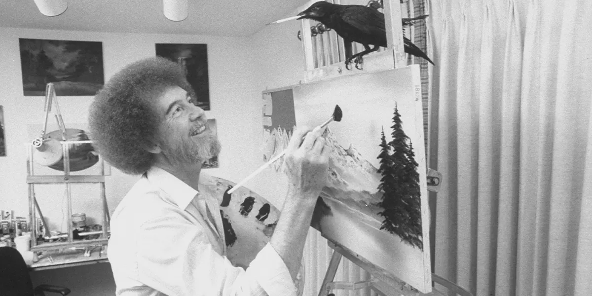 Television art teacher and artist Bob Ross paints one of his landscapes while his pet crow sits on the easel in his studio with a paintbrush in its beak. (Photo: Acey Harper/Getty Images) (detail)