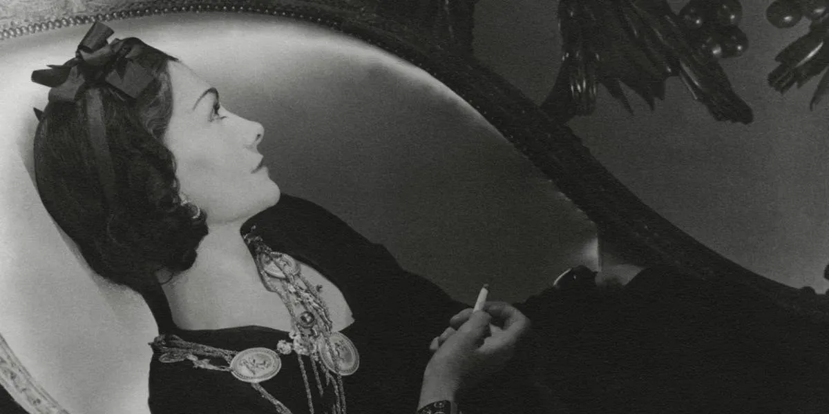 La créatrice de mode française Gabrielle Coco Chanel, de profil, allongée sur une chaise longue avec sa cigarette, portant une robe sombre avec des colliers médaillons dorés et un ruban dans les cheveux ; photographiée à Paris en 1937 (détail), image © Photo par Horst P. Horst / Conde Nast via Getty Images